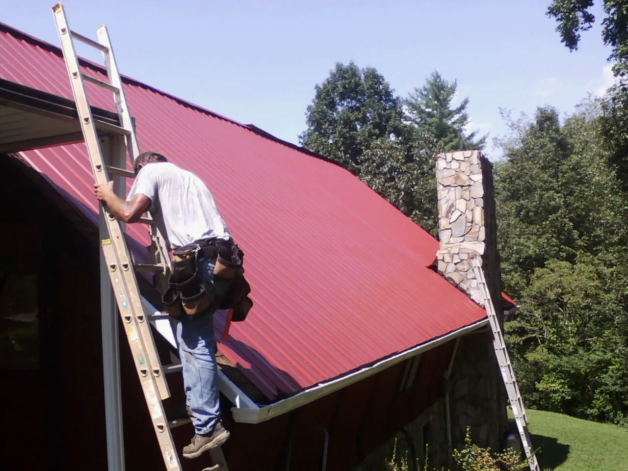 Roofer on a ladder beside a red metal roof and stone chimney — Soffit & Fascia Repair by Roofing Brothers in Notre Dame